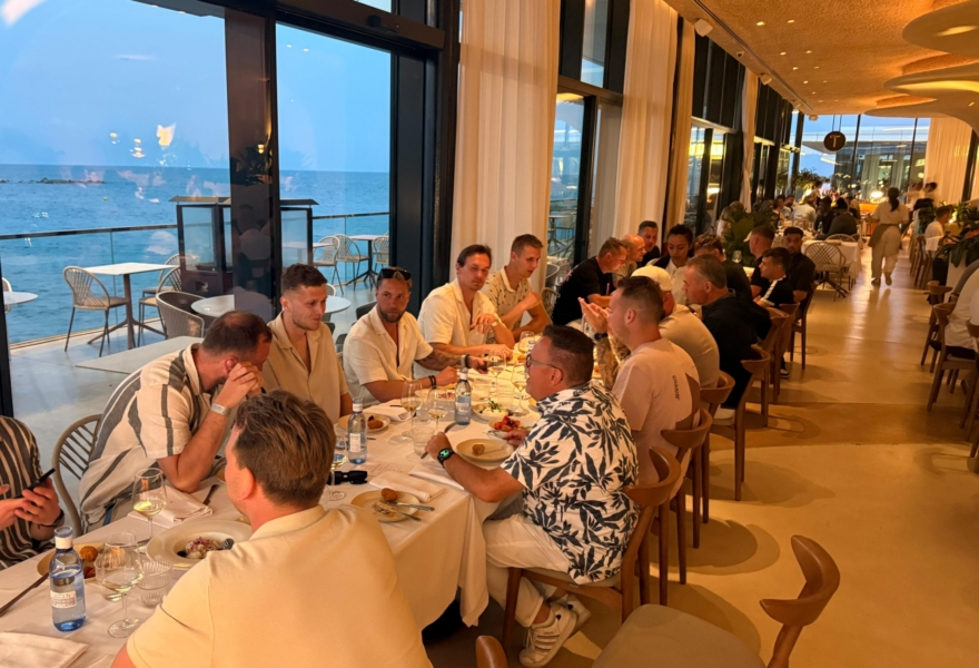 Grote groep collega’s aan een lange eettafel tijdens een gezamenlijk diner in een restaurant met uitzicht op zee in Barcelona.