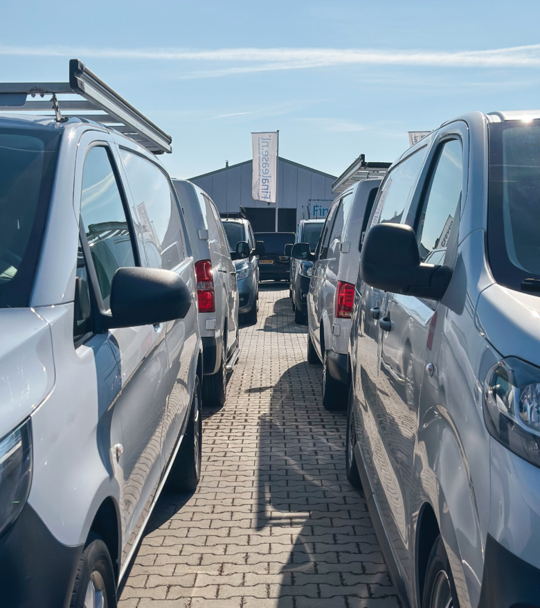 Rij bedrijfswagens die strak naast elkaar geparkeerd staan op het terrein van Greven Automotive, met een loods en vlaggen op de achtergrond.