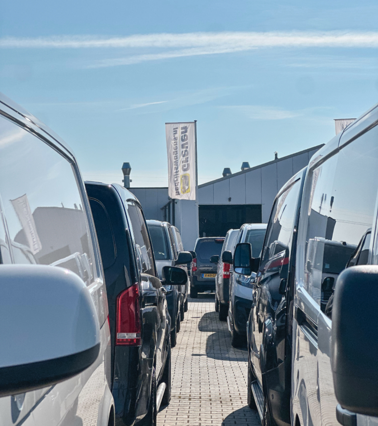 Rij bedrijfswagens die dicht naast elkaar geparkeerd staan op het terrein van Greven Automotive, met een Greven‑vlag en loods op de achtergrond.