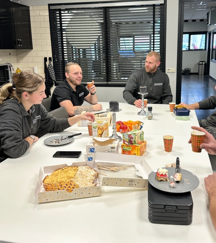 Personeel zit samen aan een tafel met koffie en gebak tijdens een pauze op kantoor.