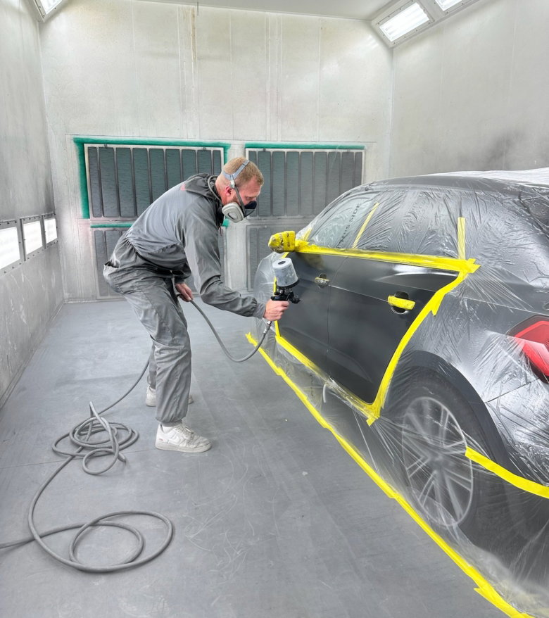 Medewerker die de zijkant van een auto spuit in een professionele spuitcabine.