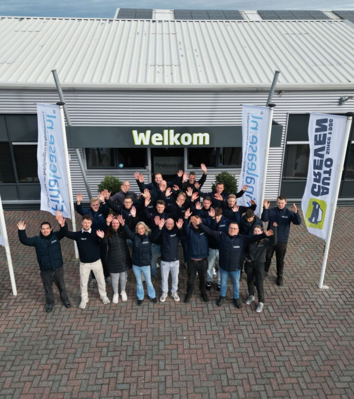 Groepsfoto van medewerkers van Greven Automotive die buiten voor Greven Automotive staan met “Welkom” boven de ingang, terwijl ze lachend en met hun handen in de lucht naar de camera zwaaien.